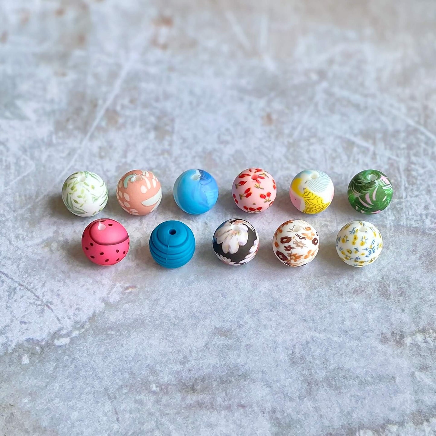 11 silicone balls designed to be used on the ends of knitting needles to stop stitches falling off are lined up on a plastered surface. They are brightly coloured.