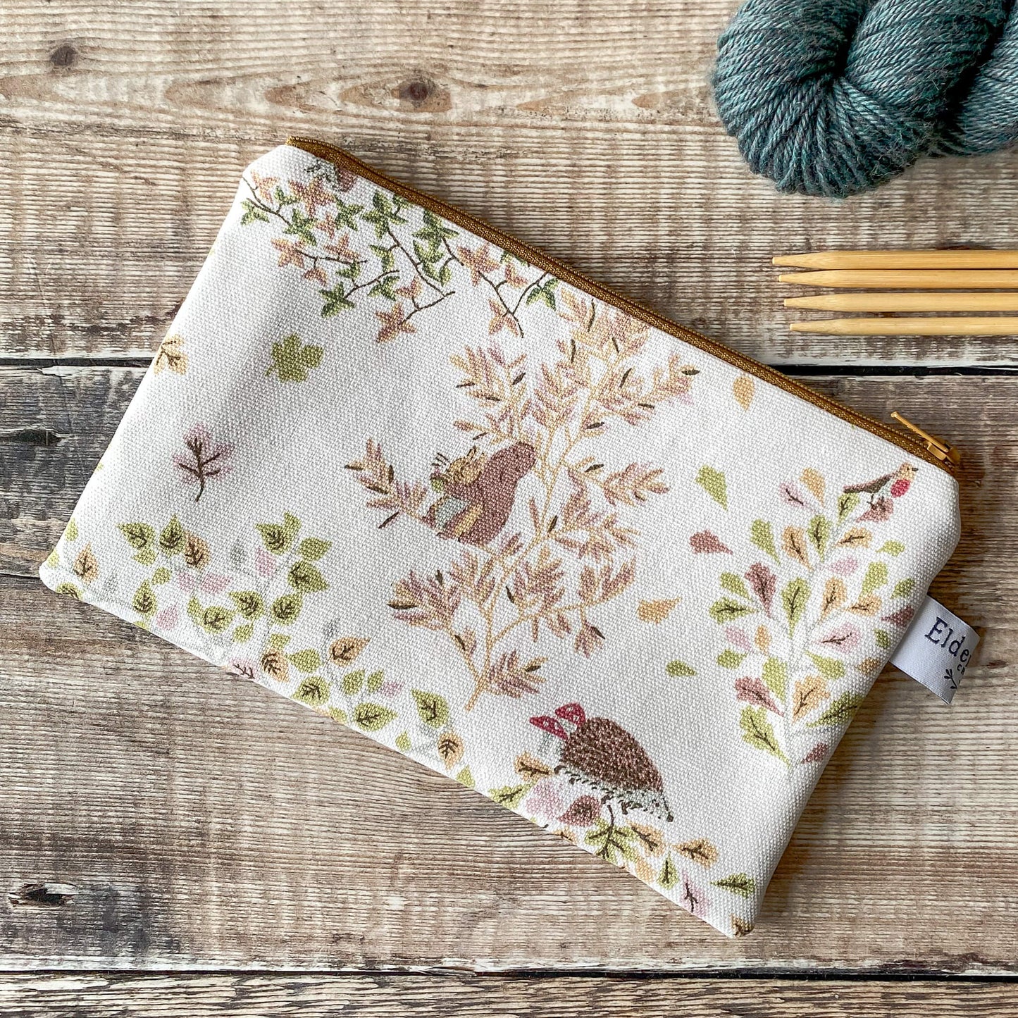 ChatGPT
The image features a small zippered pouch with a woodland pattern featuring leaves and hedgehogs. The pouch is accompanied by several skeins of yarn and knitting needles, which are placed on a wooden surface. The pouch appears to be designed for storing knitting notions, and its zipper and fabric design are visible in detail.