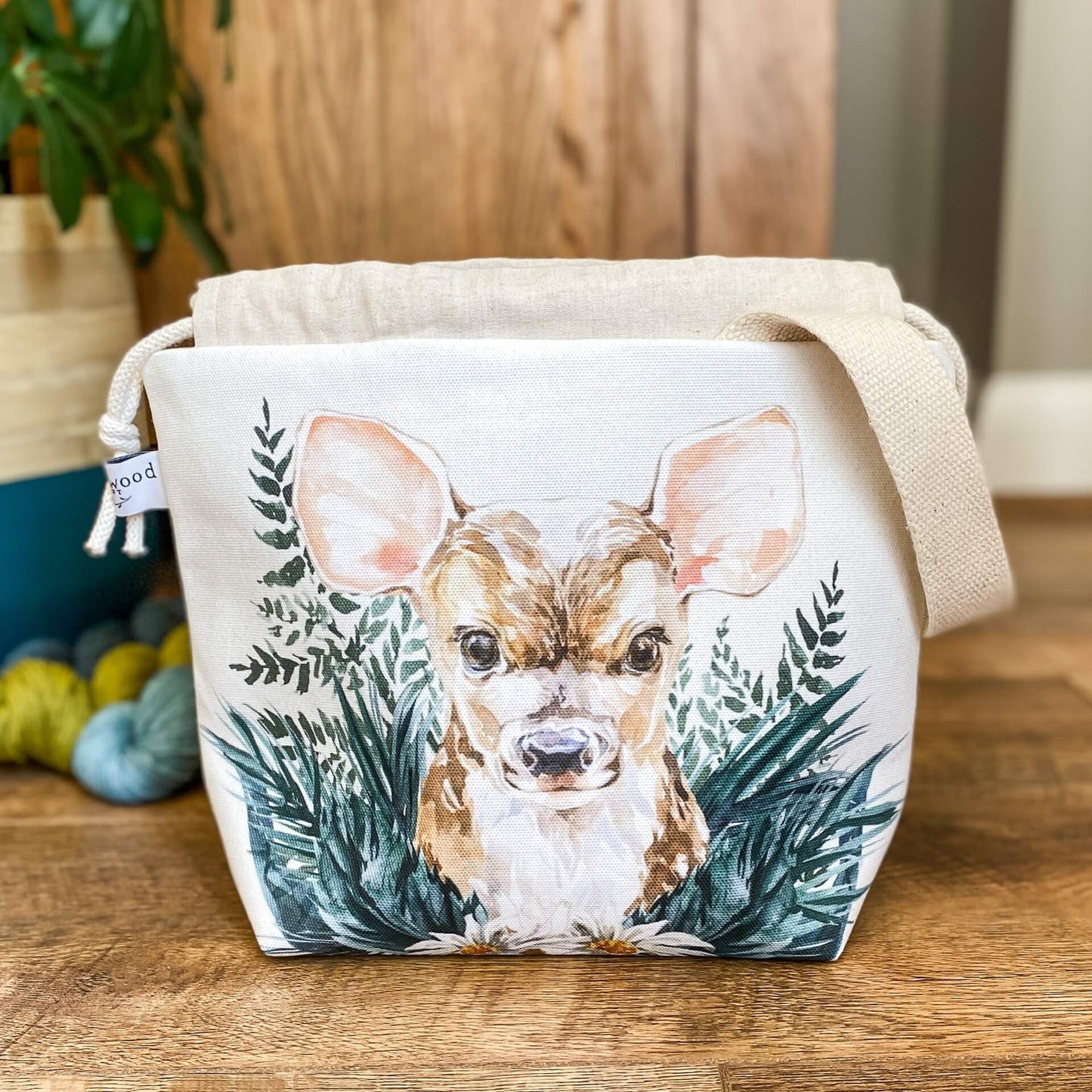 A project bag for knitting is seen on a wooden floor. The bag is a drawstring bag and on the fabric is a baby deer with big ears sitting in amongst greenery. Behind the bag can be seen a large green houseplant and some yarn on the floor. The top of the drawstring is open.