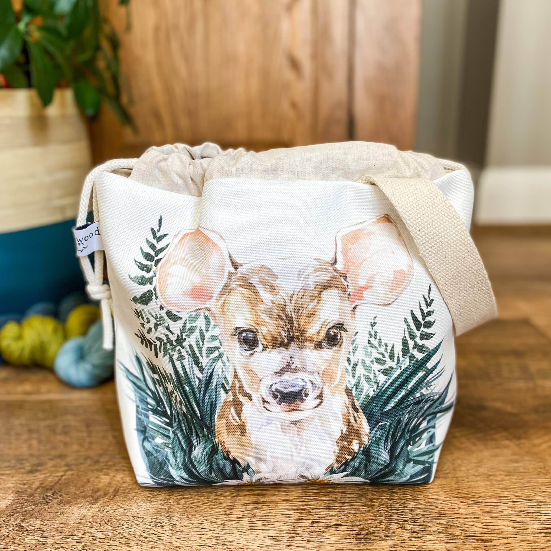 A project bag for knitting is seen on a wooden floor. The bag is a drawstring bag and on the fabric is a baby deer with big ears sitting in amongst greenery with some yarn and a house plant in the background.
