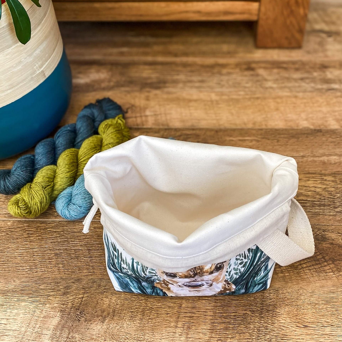 An open project bag for knitting and crochet projects sits wide open on a wooden floor allowing the user to see the plain canvas lining. Next to the bag are three skeins of yarn and a house plant in a pot.