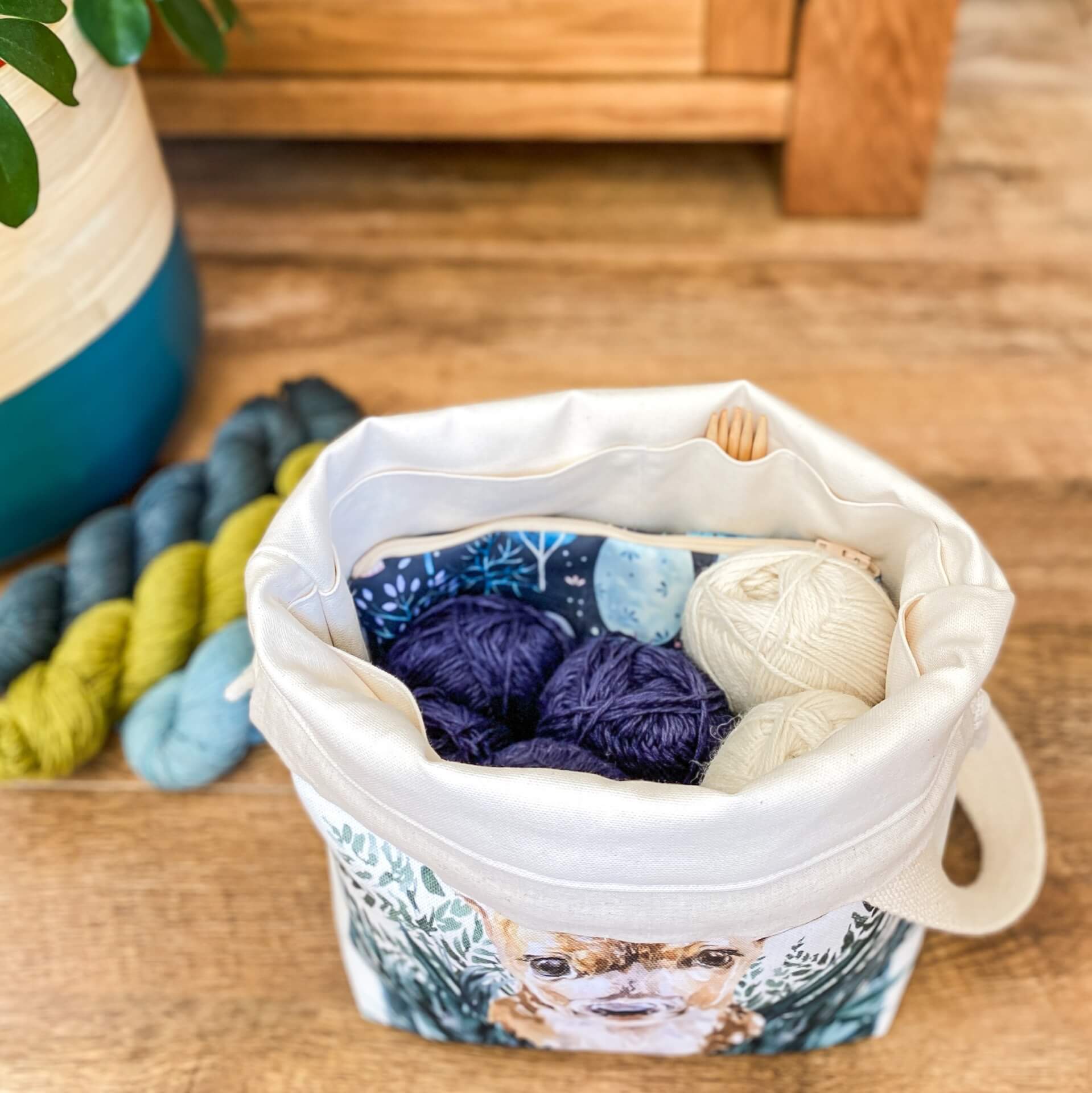 The inside of a knitting project bag is shown full of skeins of yarn and with a notions pouch tucked in. Also shown in one of the pockets are some wooden double point knitting needles. The bag sits on a floor next to three skeins of yarn and a house plant.