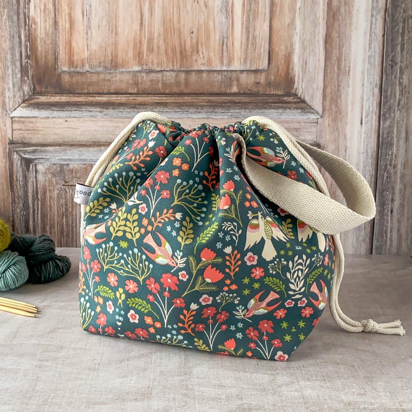 A medium-sized knitting project bag with a vibrant floral and bird design in shades of red, orange, and teal against a dark green background. The bag has fabric handles and a drawstring closure. Nearby are skeins of yarn and knitting needles on a wooden surface.