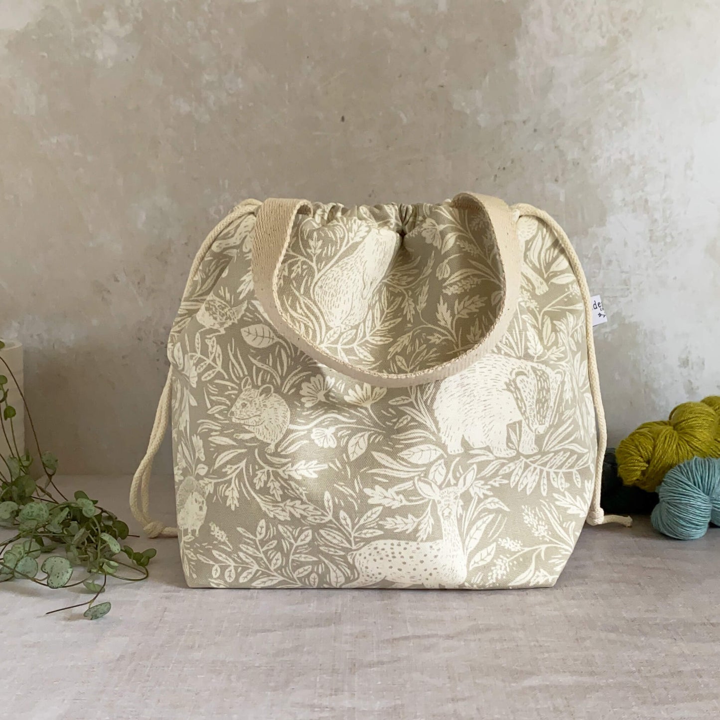 A large sized handmade project bag in a linen coloured countryside themed fabric. A drawstring closure at the top, and a sturdy handle, ideal for carrying knitting or crochet supplies and projects. Ideal for organising large knitting and crochet projects such as garments and blankets. The interior of the bag is made from a cream canvas and has lots of storage pockets. The bag sits in front of three skeins of yarn and a pot plant.