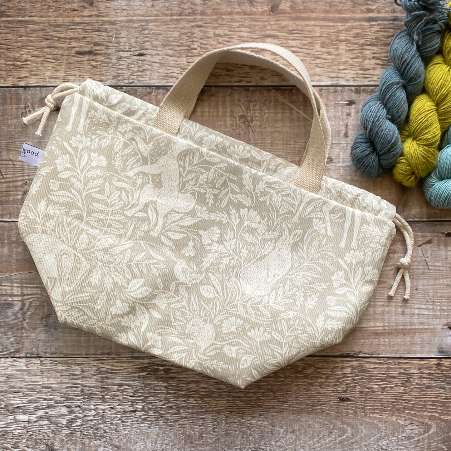 A large sized handmade project bag in a linen coloured countryside themed fabric, featuring a drawstring closure and sturdy handle, perfect for carrying knitting or crochet supplies. The bag is displayed next to three skeins of yarn and some wooden knitting needles.