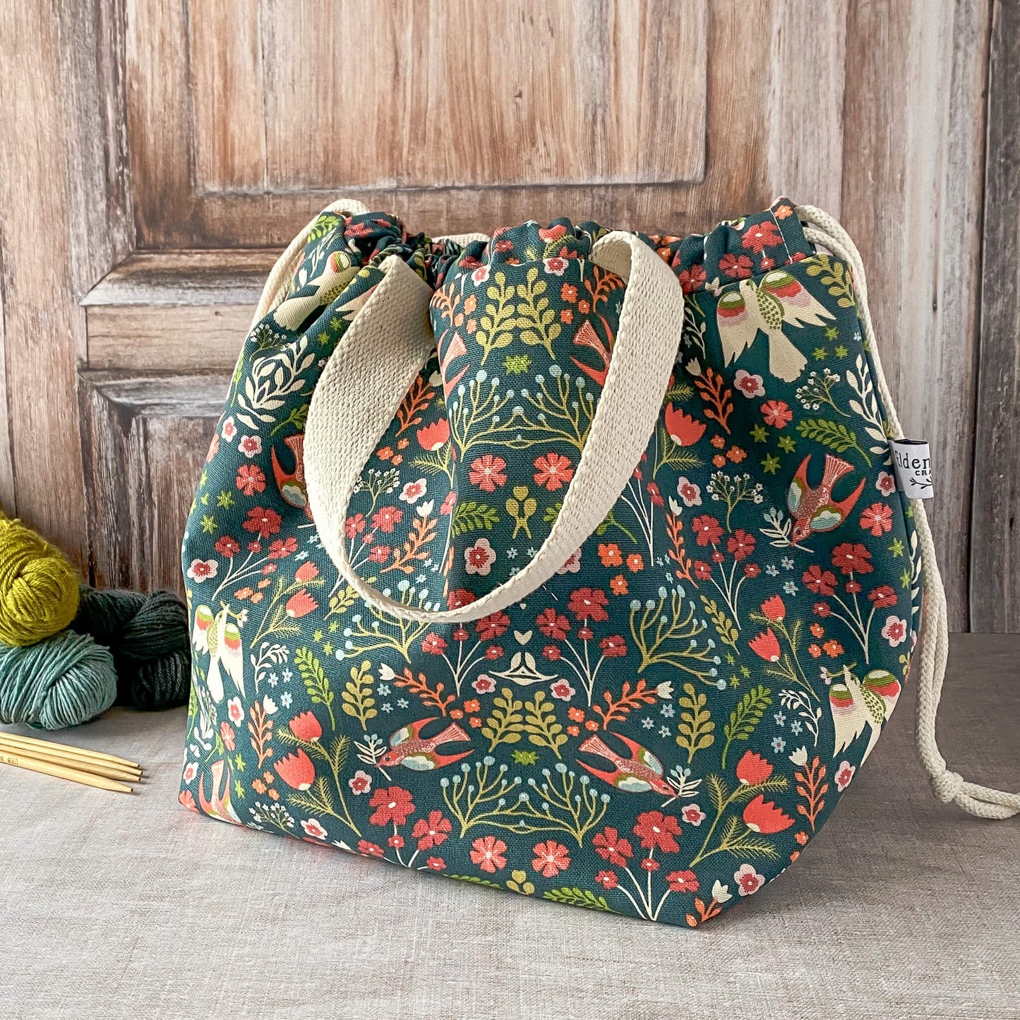 This image shows a large knitting project bag with a floral pattern, featuring bright flowers and birds in red, orange, and teal against a dark green background. The bag is cinched at the top with a drawstring, and it has sturdy light-colored fabric handles. The bag is placed on a neutral surface, with knitting needles and yarn in the background.