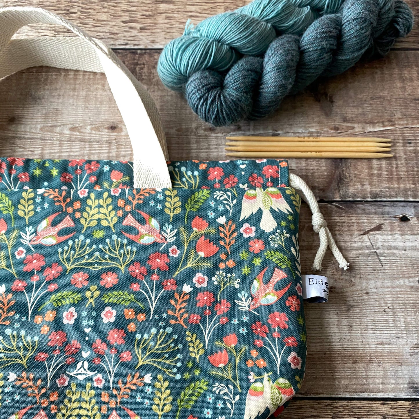 This image shows a large knitting project bag with a colorful floral design featuring birds and flowers in shades of red, orange, teal, and green against a dark background. The bag has sturdy cream-colored handles and a drawstring closure. Next to the bag, there are skeins of yarn and a set of knitting needles on a wooden surface. The brand label is visible on the side of the bag.
