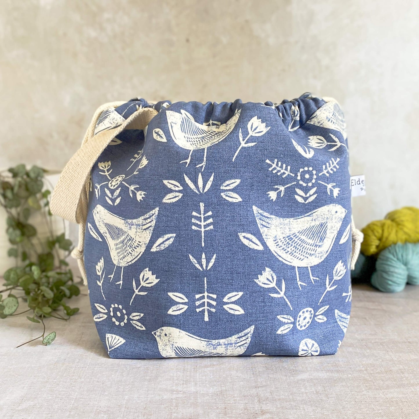 A medium sized handmade project bag in a Scandi inspired blue bird-themed fabric. A drawstring closure at the top, and a sturdy handle, ideal for carrying knitting or crochet supplies and projects. Ideal for organising shawl, scarf and lightweight garment projects. The interior of the bag is made from a cream canvas and has lots of storage pockets. The bag sits in front of three skeins of yarn and a pot plant.