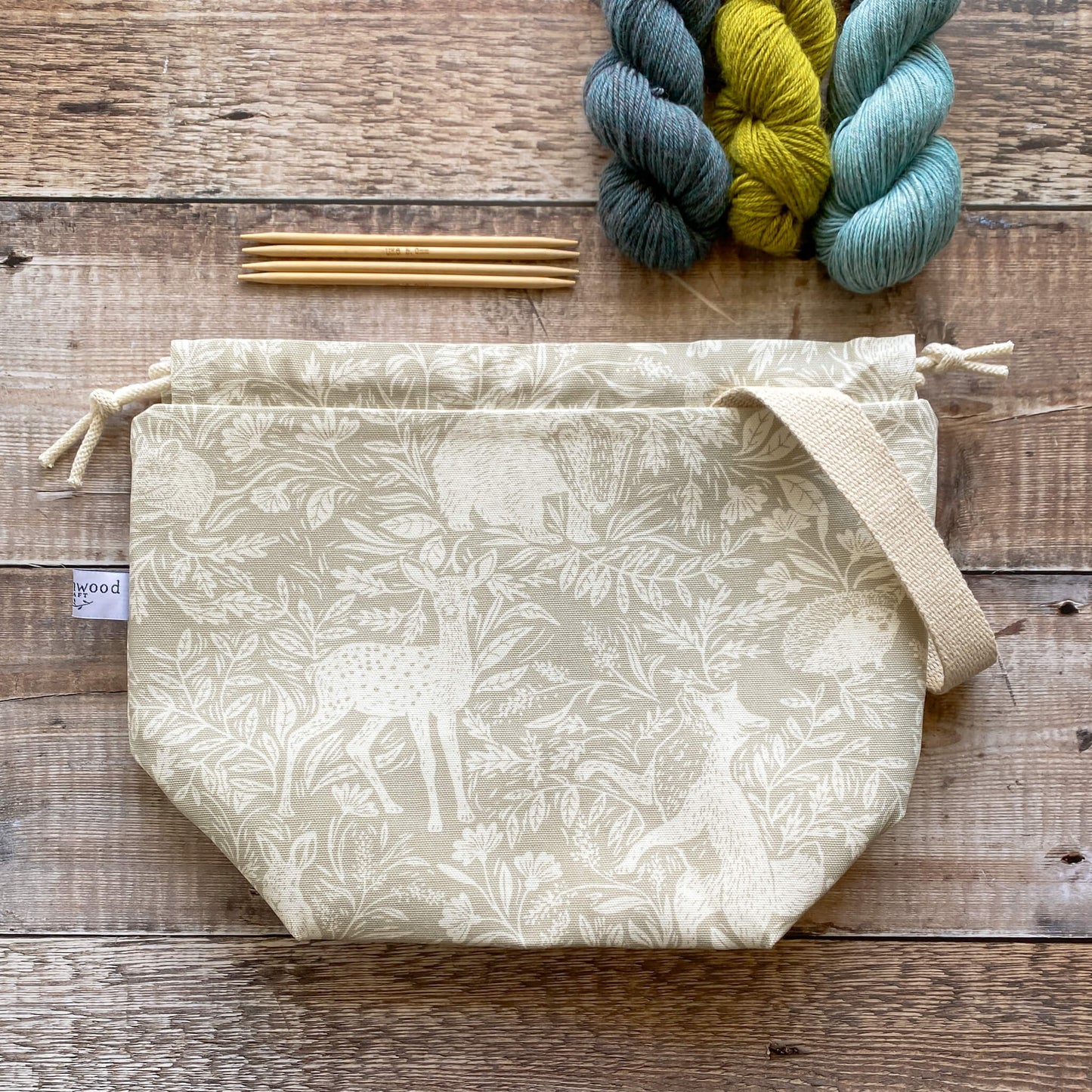 A medium sized handmade project bag in a linen coloured countryside fabric, featuring a drawstring closure and sturdy handle, perfect for carrying knitting or crochet supplies. The bag is displayed next to three skeins of yarn and some wooden knitting needles.