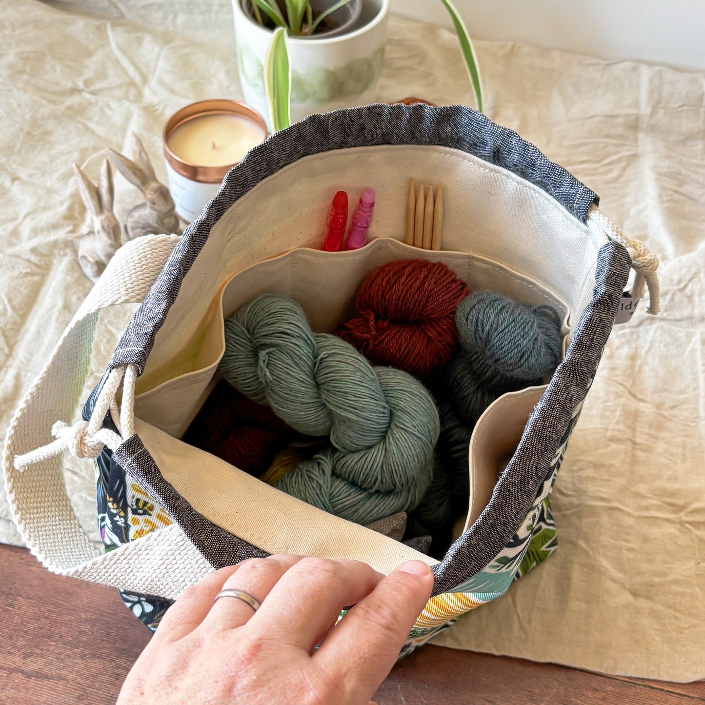 A fabric and canvas storage tote bag filled with skeins of yarn and knitting tools, held open by a person's hand on a beige fabric surface.