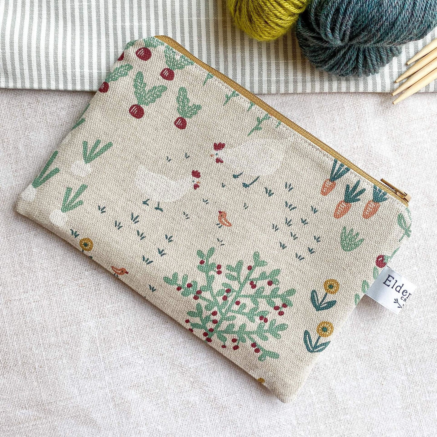 A small zipped pouch intended to be used for knitting notions lies on a table top next to two skeins of yarn and some wooden knitting needles. On the front of the pouch we can see some chickens, a bird, some beetroot and carrots, all printed in a whimsical style.