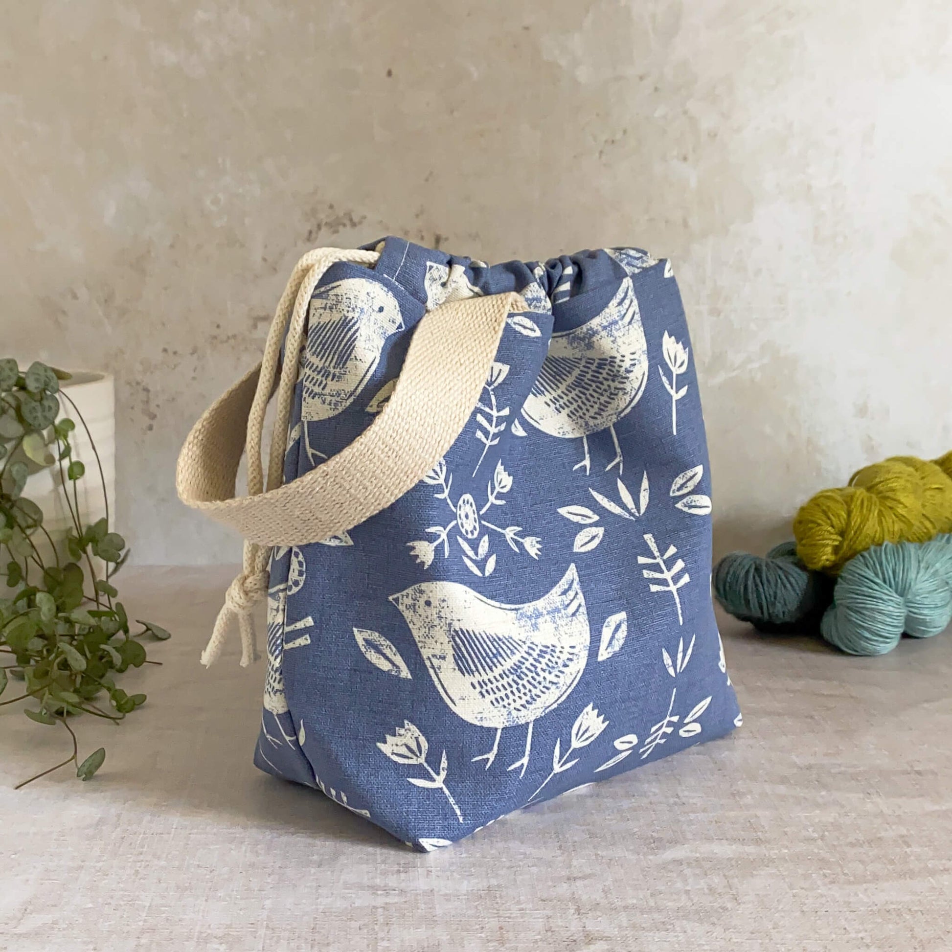 A side on view of a compact handmade project bag in Scandi inspired blue bird themed fabric, featuring a drawstring closure and sturdy handle, perfect for organizing knitting and crochet supplies. Displayed with three skeins of yarn and a pot plant in the background.
