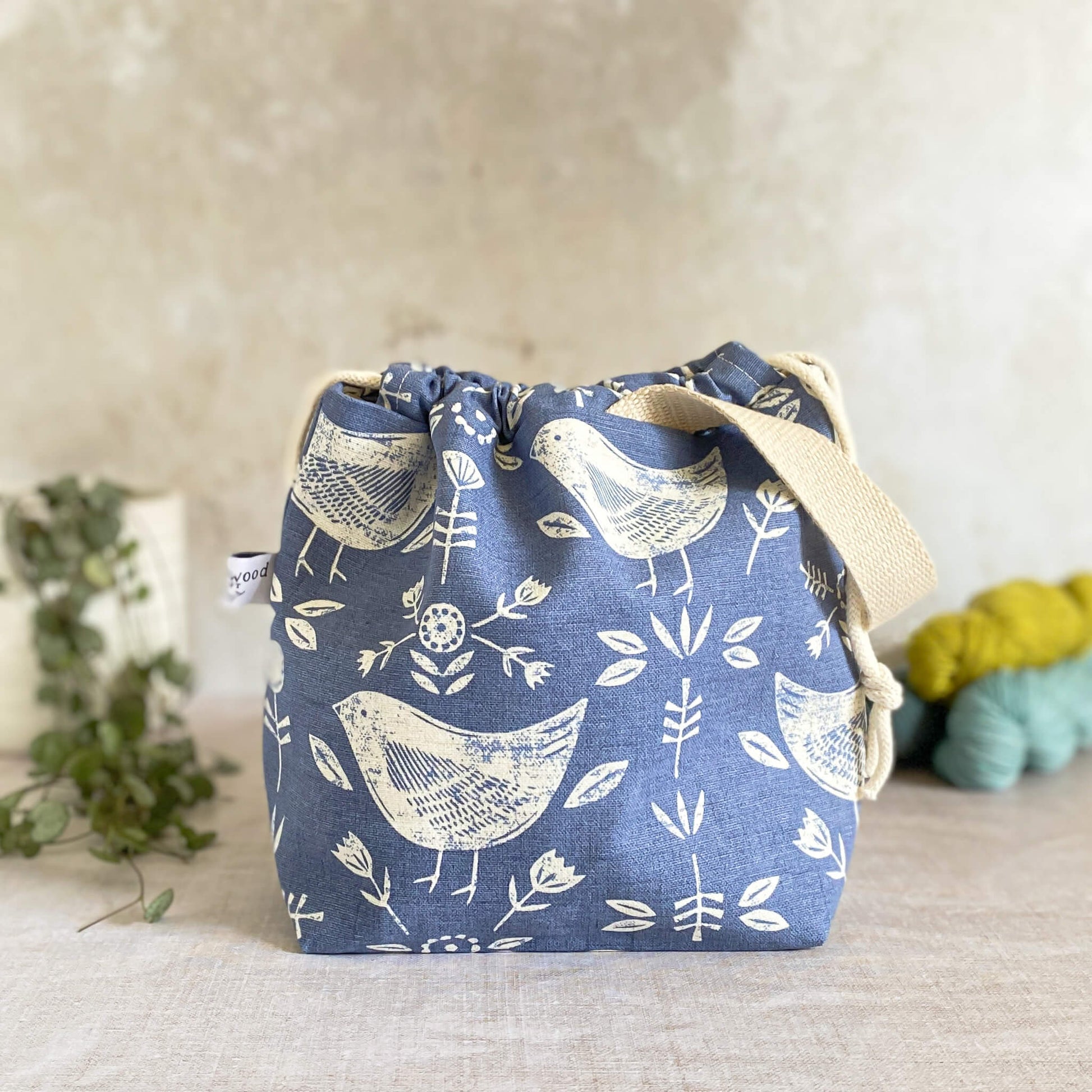 Small handmade project bag crafted from Scandinavian-inspired blue bird-themed fabric, featuring a convenient drawstring closure and sturdy handle. Perfect for keeping knitting and crochet supplies organized, complemented by three skeins of yarn and a potted plant in the backdrop.