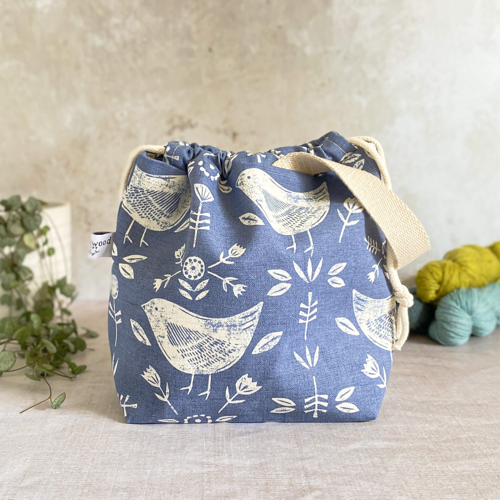 Handmade project bag in charming Scandi blue bird fabric, designed with a practical drawstring closure and durable handle. Ideal for neatly organizing knitting and crochet essentials, showcased with three skeins of yarn and a potted plant in the background.