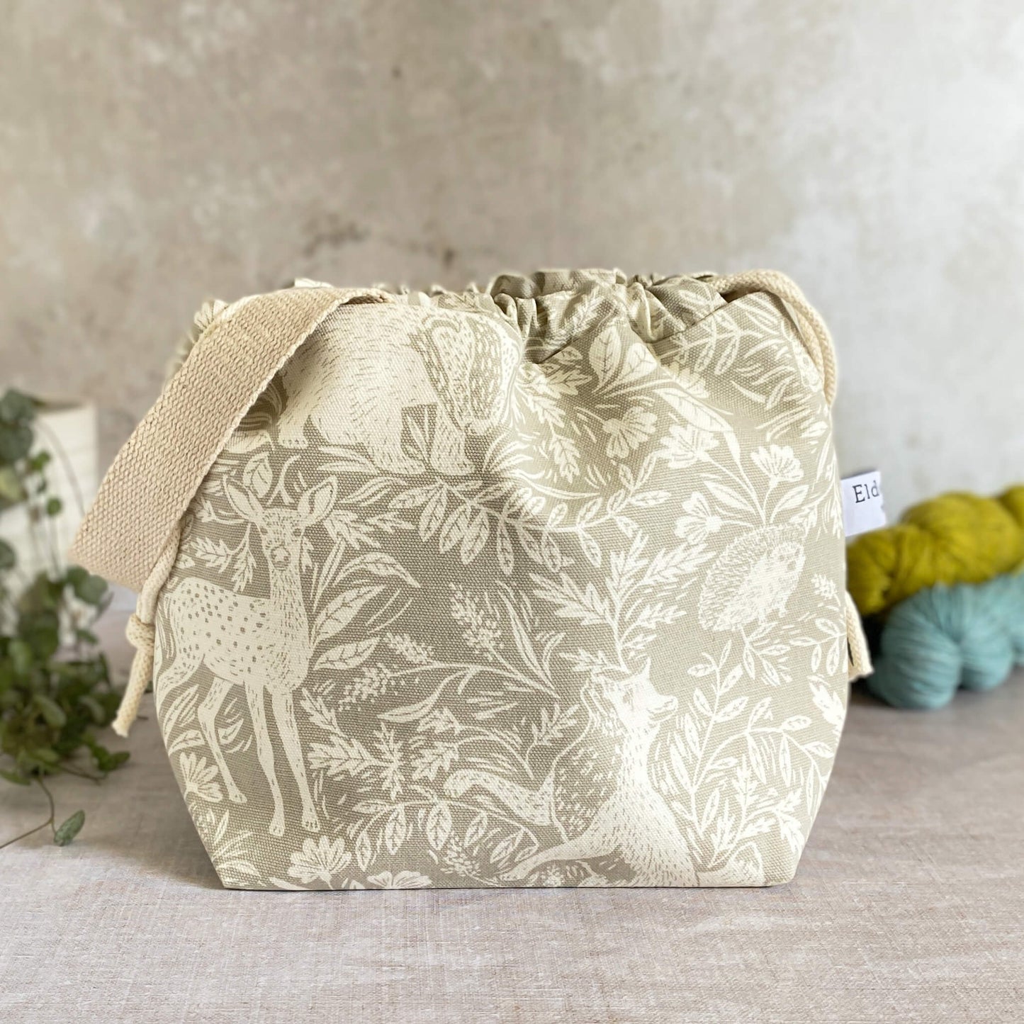 A small sized handmade project bag in a countryside fabric. A drawstring closure at the top, and a sturdy handle, ideal for carrying knitting or crochet supplies and projects. The bag sits in front of three skeins of yarn and a pot plant.