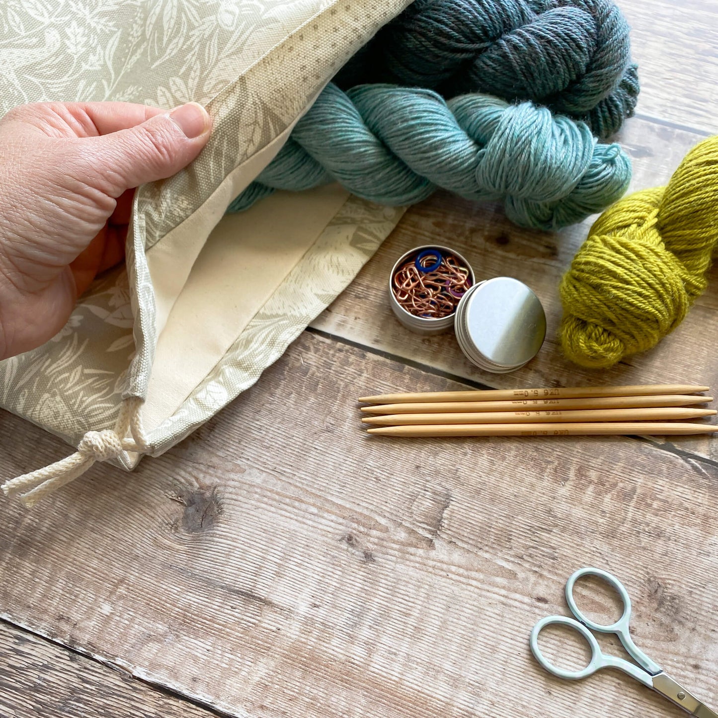 An view inside a linen coloured countryside themed handmade project bag. Two skeins of yarn poke out of the bag which is being held open by a pale skinned hand. Next to the bag is some more yarn, some stitch markers, scissors and wooden knitting needles.