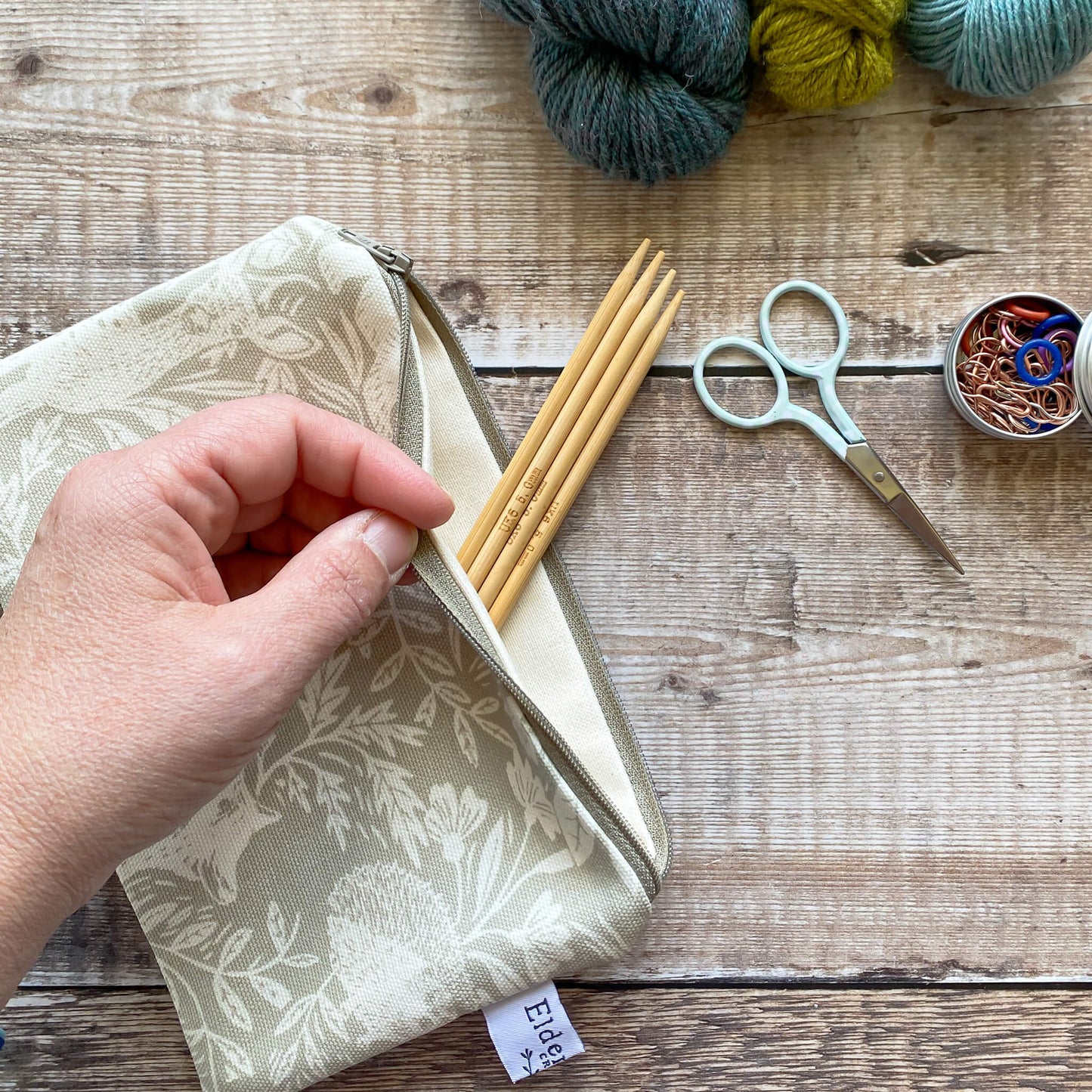 A small zipped notions pouch is held open by a pale skinned hand revealing the cream coloured canvas lining. Four wooden knitting needles poke out of the bag. Next to the bag on a wooden table are three skeins of yarn, scissors and some stitch markers.