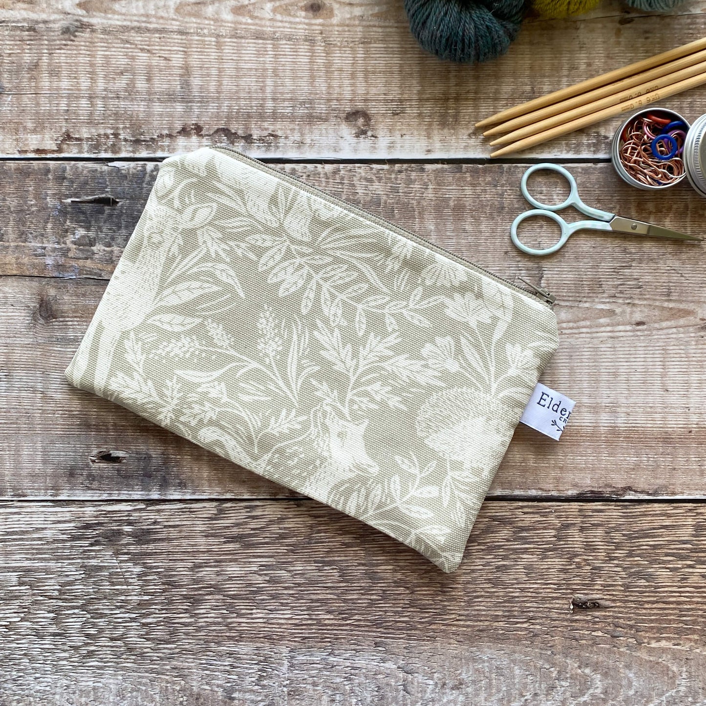 A small hand made zipped pouch featuring a linen coloured countryside themed fabric lies on a wooden table. The pouch is intended to be used to hold knitting and crochet notions. Next to the pouch are some scissors, knitting needles and stitch markers.