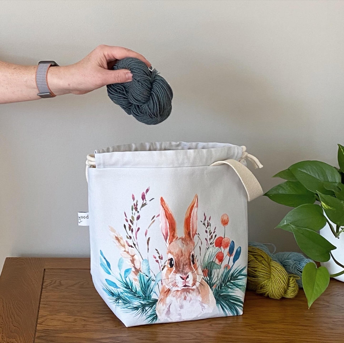 A handmade project bag from Eldenwood Craft is sitting a wooden table next to two skeins of yarn and a plant. The project bag features a watercolour hare surrounded by leaves and shoots and a skein of blue yarn is being placed into the bag.