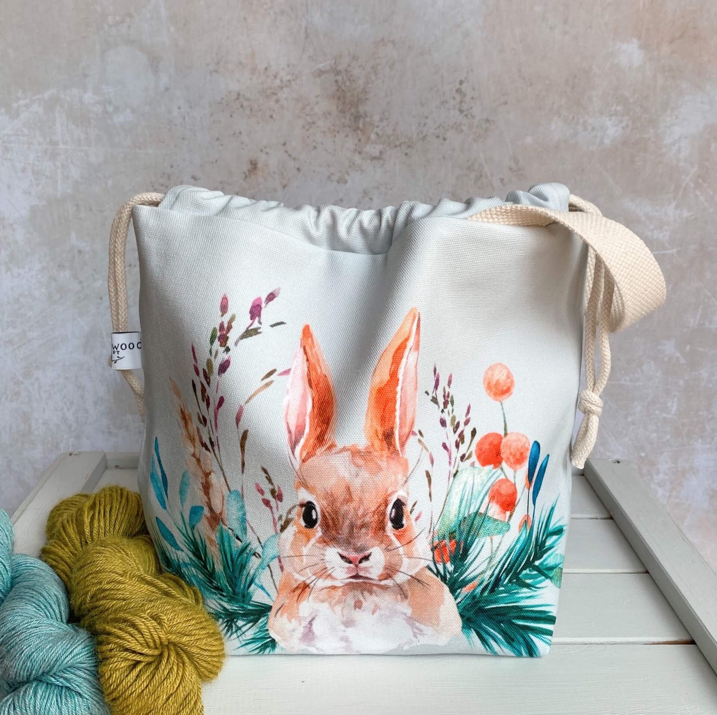 A knitting project bag handmade by Eldenwood Craft featuring a watercolour hare print. The project bag is sitting on a bench next to two skeins of yarn.