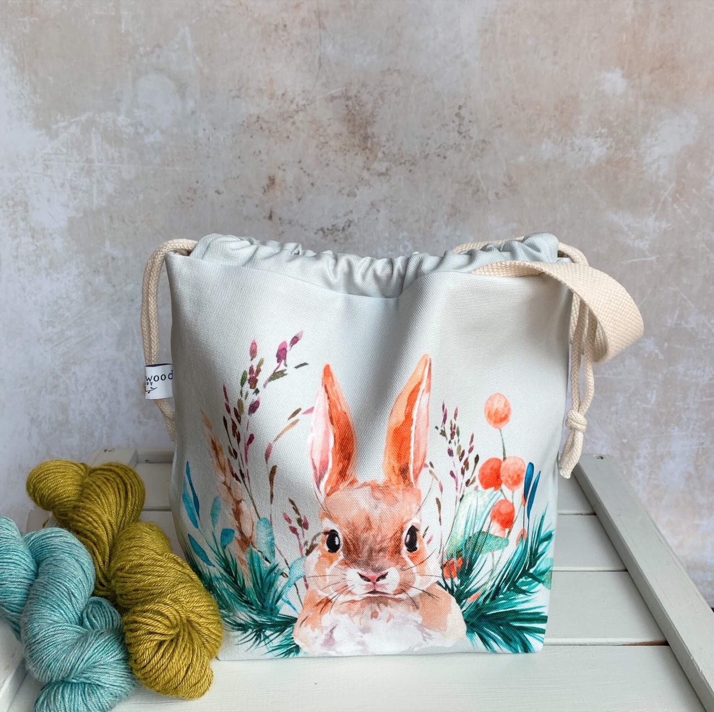 A knitting project bag handmade by Eldenwood Craft featuring a watercolour hare print. The project bag is sitting on a bench next to two skeins of yarn.