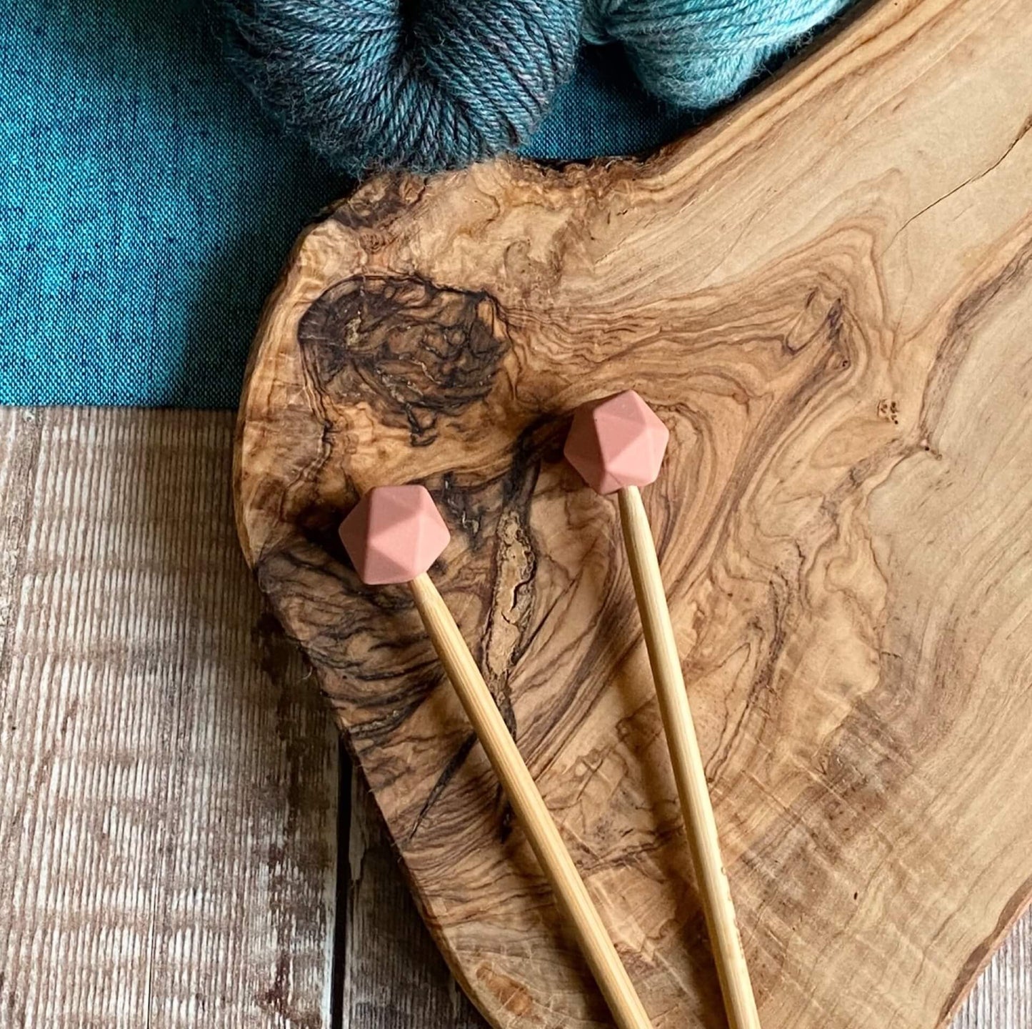 Dark pink needle protectors to stop stitches escaping from knitting needles. A pair sit on a wooden table attached to knitting needles. 