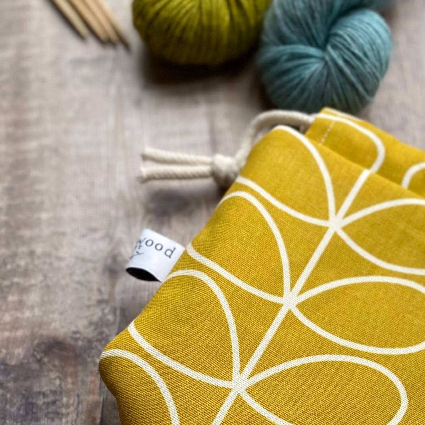 A close up of a section of a yellow project bag lying on a table next to some yarn and knitting needles. The print of the fabric is a classic Orla Kiely linear stem print and the label of the maker, Eldenwood Craft, can be seen attached to the bag.