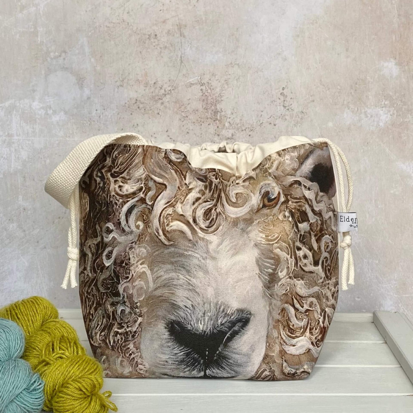 A large sheep face adorns the front of a knitting project bag. The bag has a sturdy white handle and sits on a bench next to two skeins of yarn.