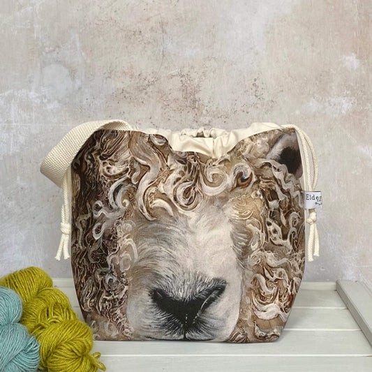 A large sheep face adorns the front of a knitting project bag. The bag has a sturdy white handle and sits on a bench next to two skeins of yarn.
