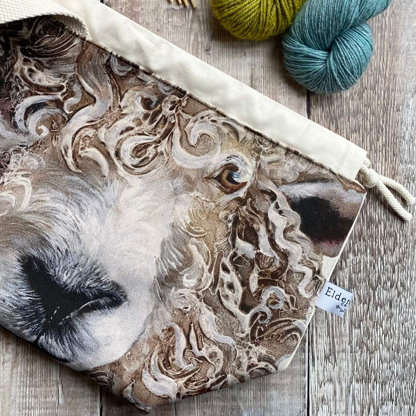 A knitting project bag for medium sized projects lies on a wooden table next to two skeins of yarn. The project bag is handmade and features a large sheep face.