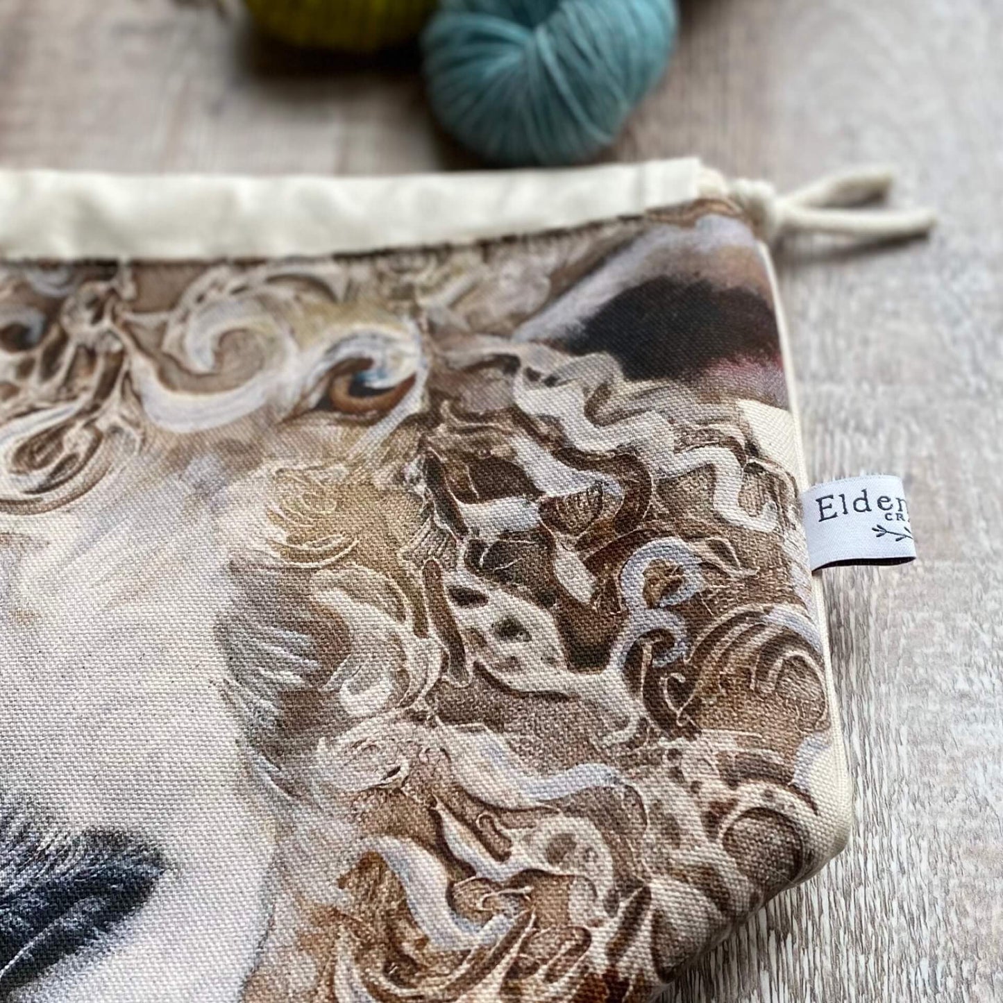 A close up of a large sheep face which is on the front of a handmade project bag sitting on a wooden table.