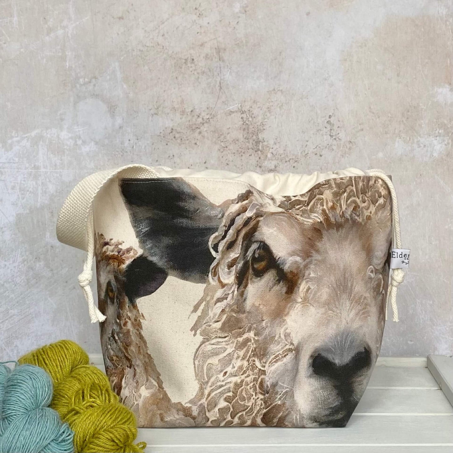 Two sheep stare out of the front of a medium sized knitting project bag that is sitting on top of a bench, next to two skeins of yarn. The project bag has a sturdy natural coloured handle.