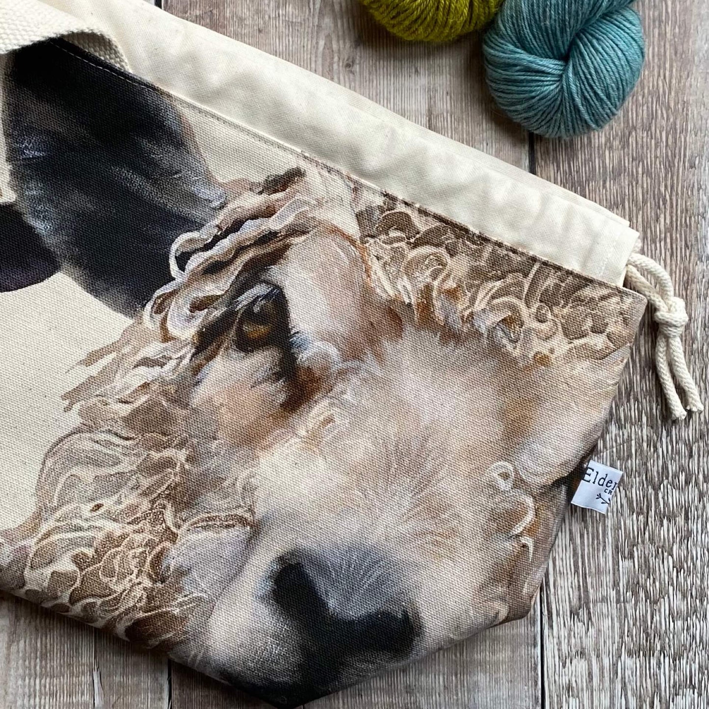 A handmade project bag with two sheep face in large scale sits on top of a wooden table next to some knitting needles and skeins of yarn.
