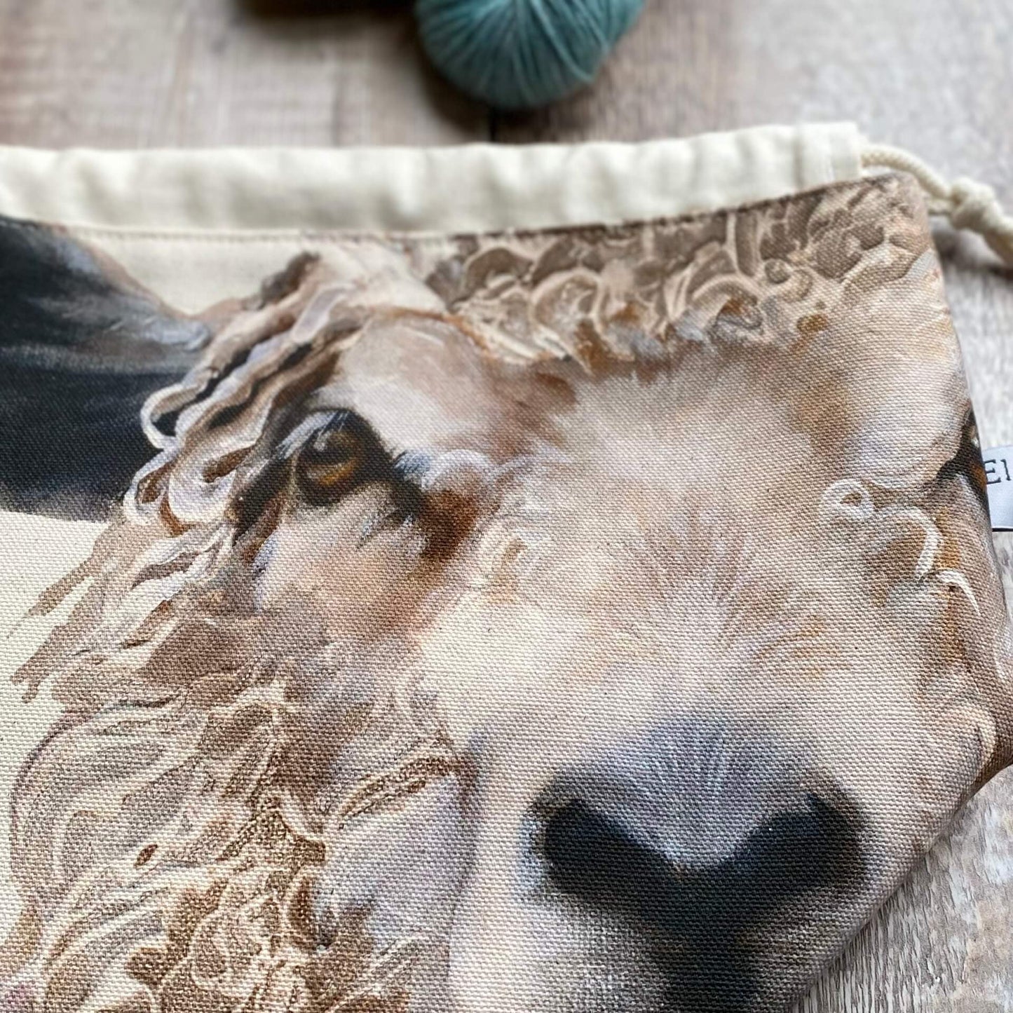A close up of a knitting project bag lying on a wooden table next to some yarn. The project bag is made using a print featuring two sheep faces.