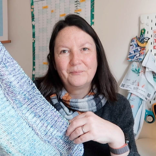 Emma, the host of the Eldenwood Craft knitting podcast, holds up a brioche shawl in her studio 
