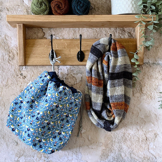 A project bag hangs from a coat peg. Next to it is a Litmus Cowl and on the shelf above are some balls of yarn and a trailing plant. 