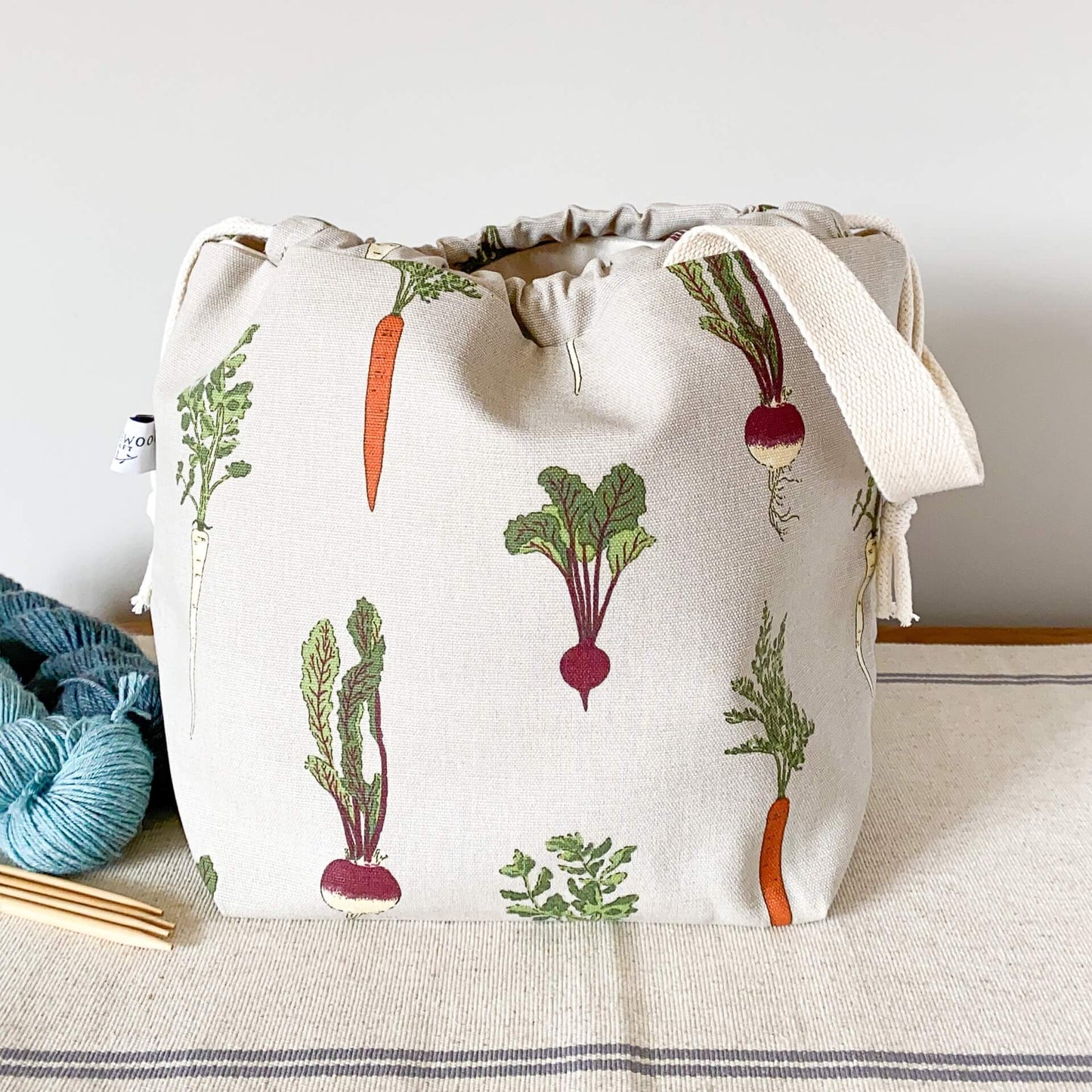 A project bag for knitting projects, made using a fabric showing off lots of home grown vegetables, sits on a table next to some skeins of yarn. 