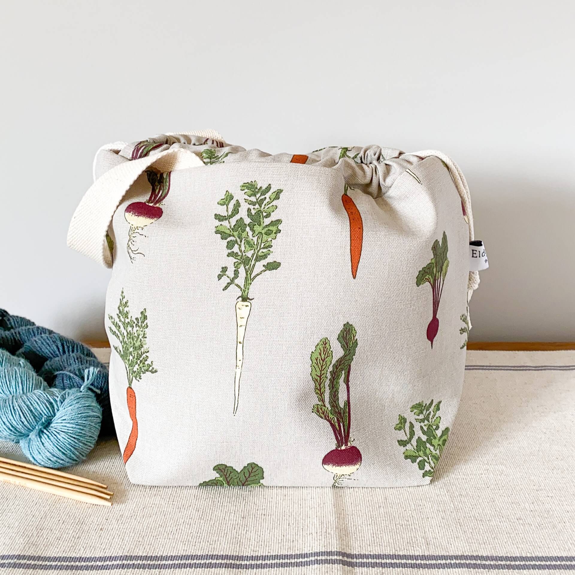 A project bag for knitting projects, made using a fabric showing off lots of home grown vegetables, sits on a table next to some skeins of yarn. The bag is pulled closed hiding its contents. 