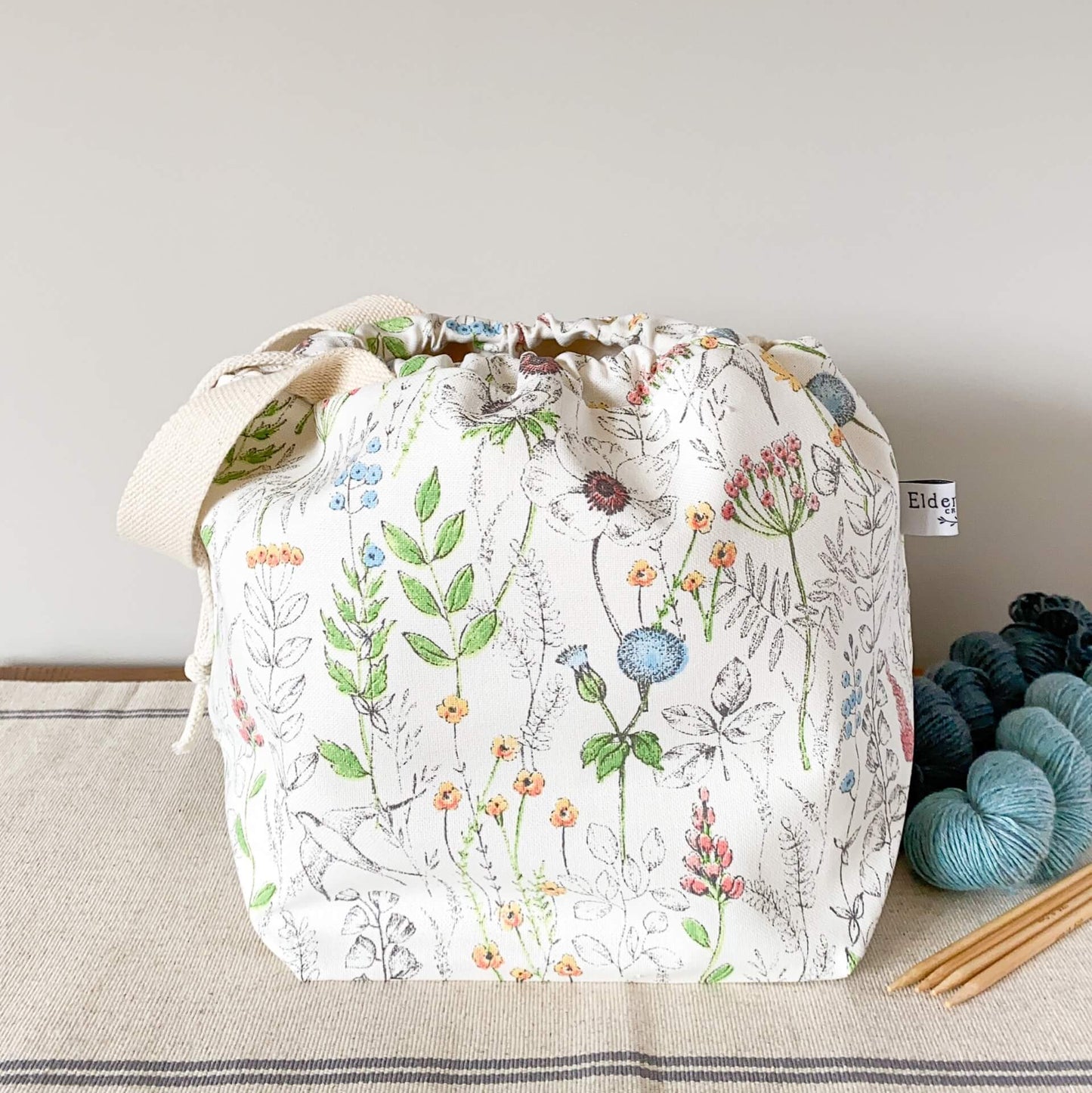 A summery project bag that features lots of wild flowers, birds and butterflies, sits on a table next to some skeins of yarn and a set of wooden knitting needles. The bag is pulled closed by a drawstring hiding its contents. 