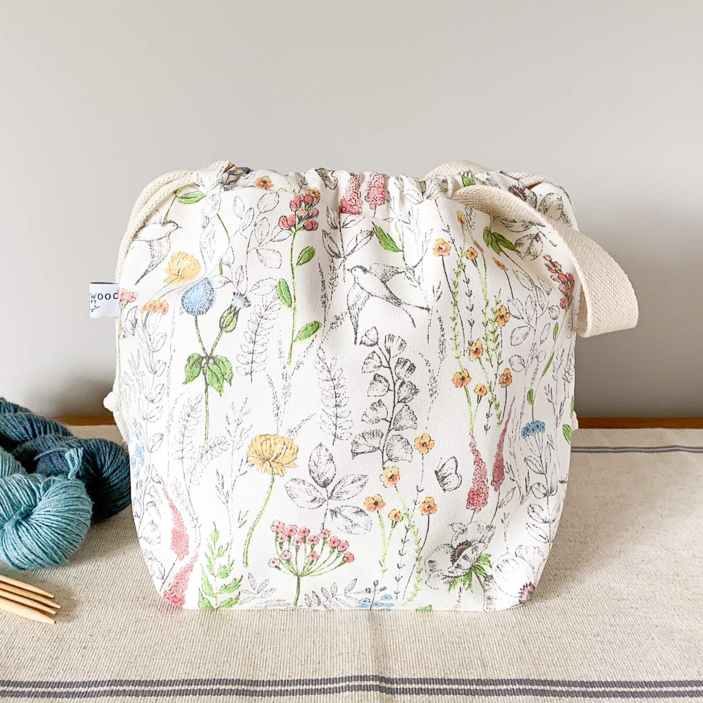 A summery project bag that features lots of wild flowers, birds and butterflies, sits on a table next to some skeins of yarn and a set of wooden knitting needles. 
