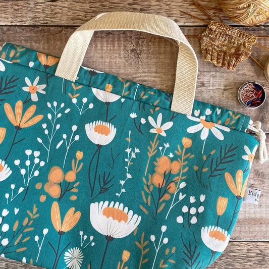 A brightly coloured floral project bag sits on a wooden table next to a knitting project and some stitch markers. The main fabric of the bag is a lovely teal colour and has lots of botanical elements printed on it. 
