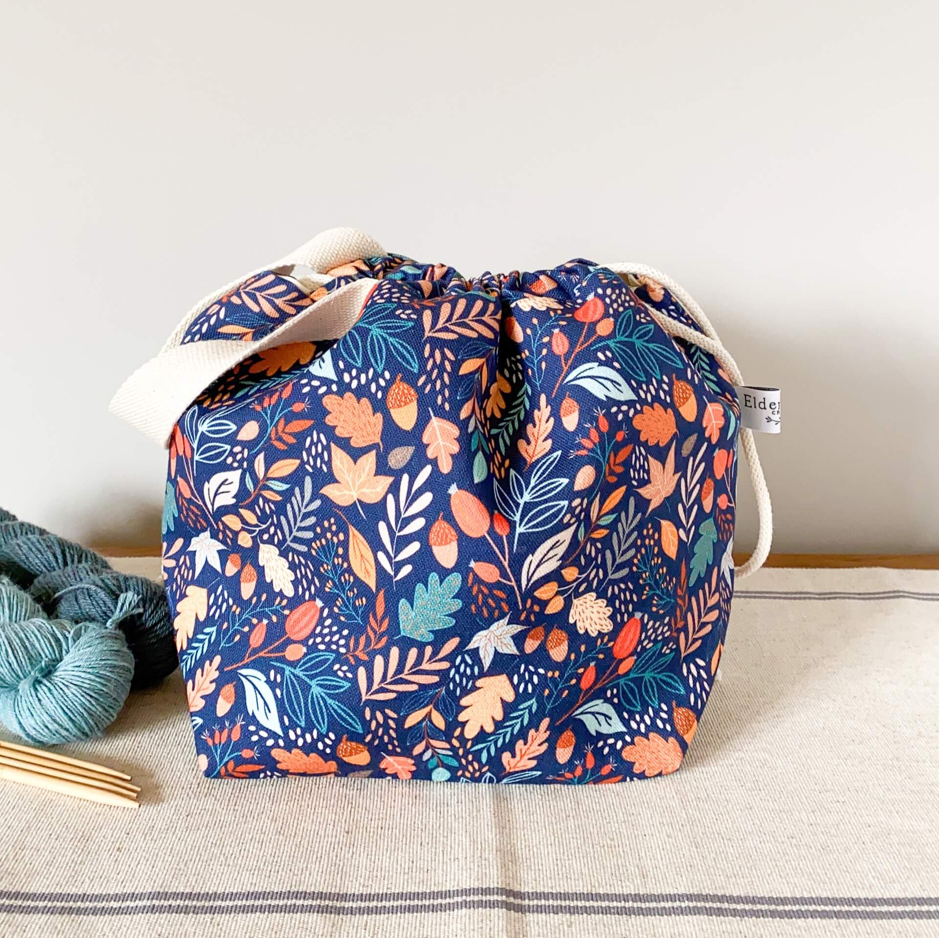 A deep blue project bag sits on a table next to two skeins of yarn. The blue fabric is printed with lots of small leaves and berries and acorns. The bag is pulled closed. 