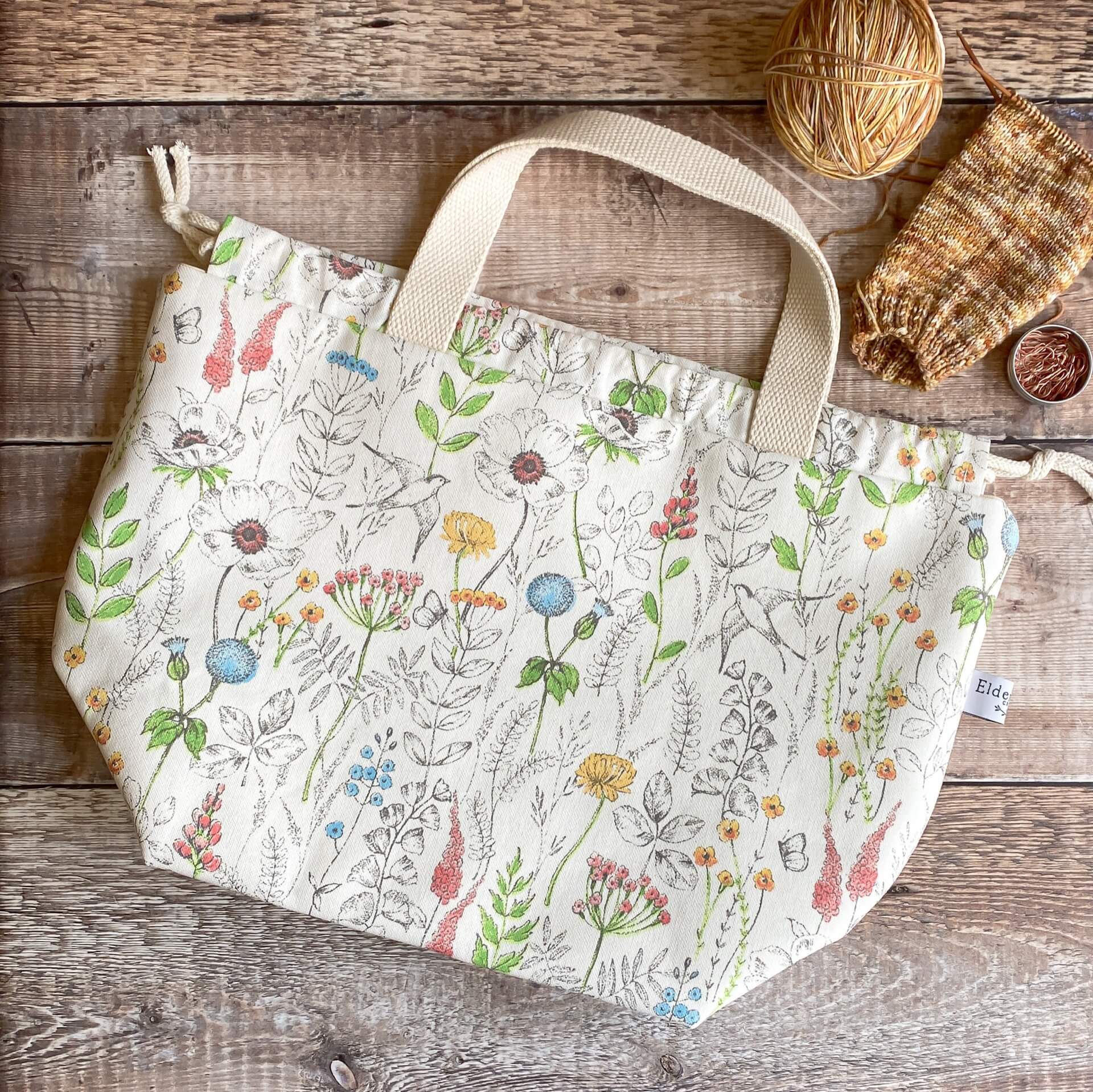 A large knitting project bag in a summery floral print sits on a wooden table next to an in progress knitting project.