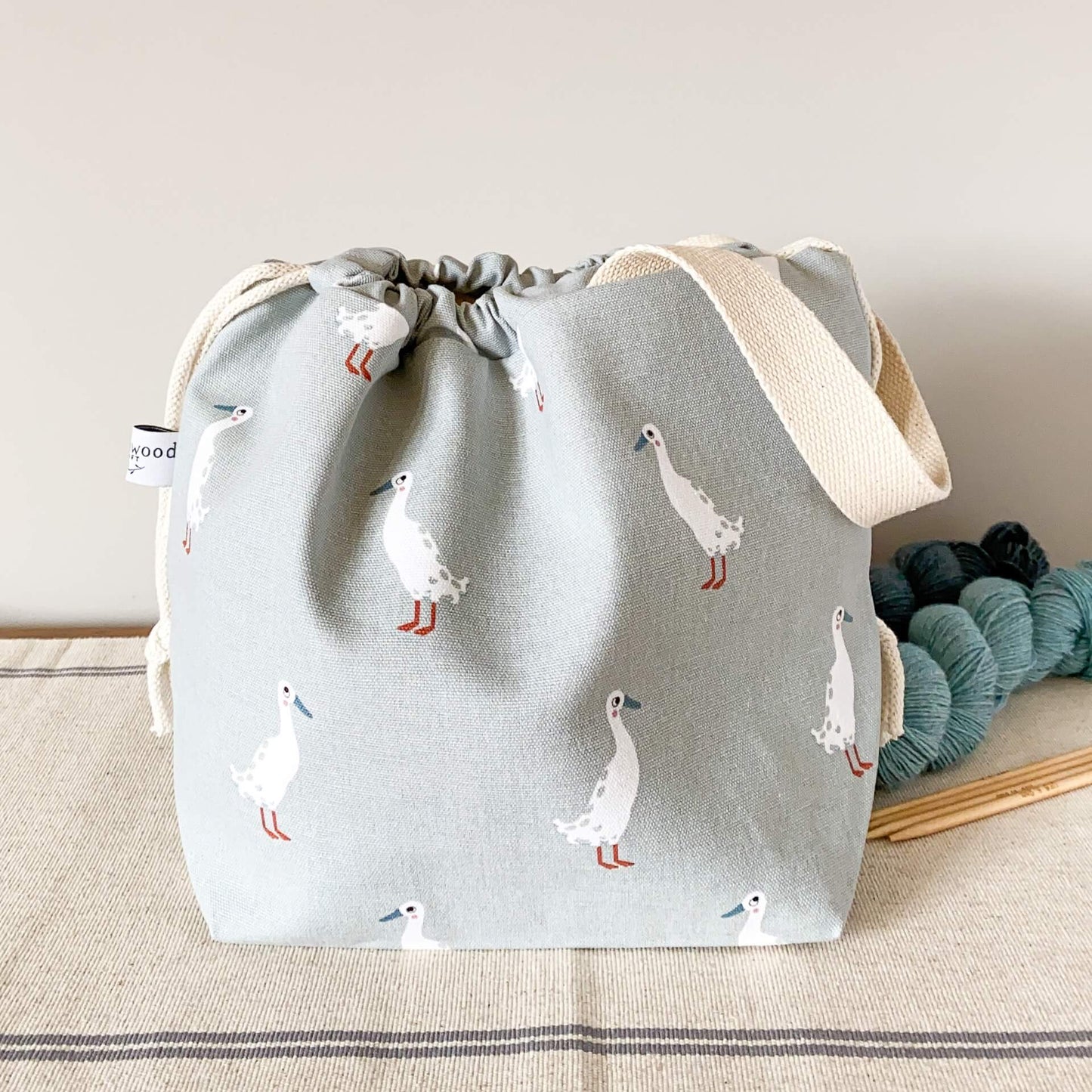 A project bag for knitting projects, made using a fabric showing lots of white runner ducks, sits on a table next to some skeins of yarn. 