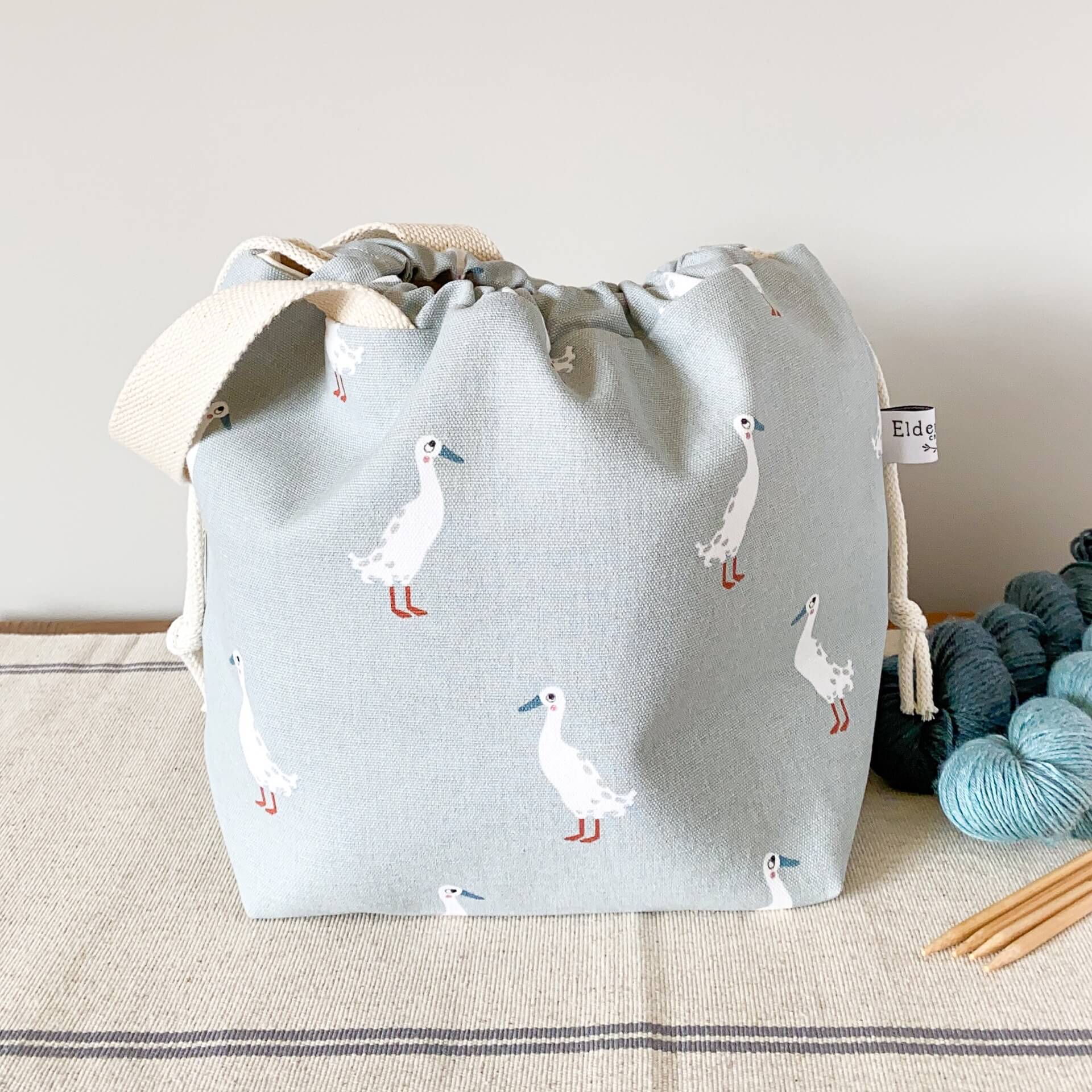 A project bag for knitting projects, made using a fabric showing lots of white runner ducks, sits on a table next to some skeins of yarn. The bag is pulled closed hiding its contents. 