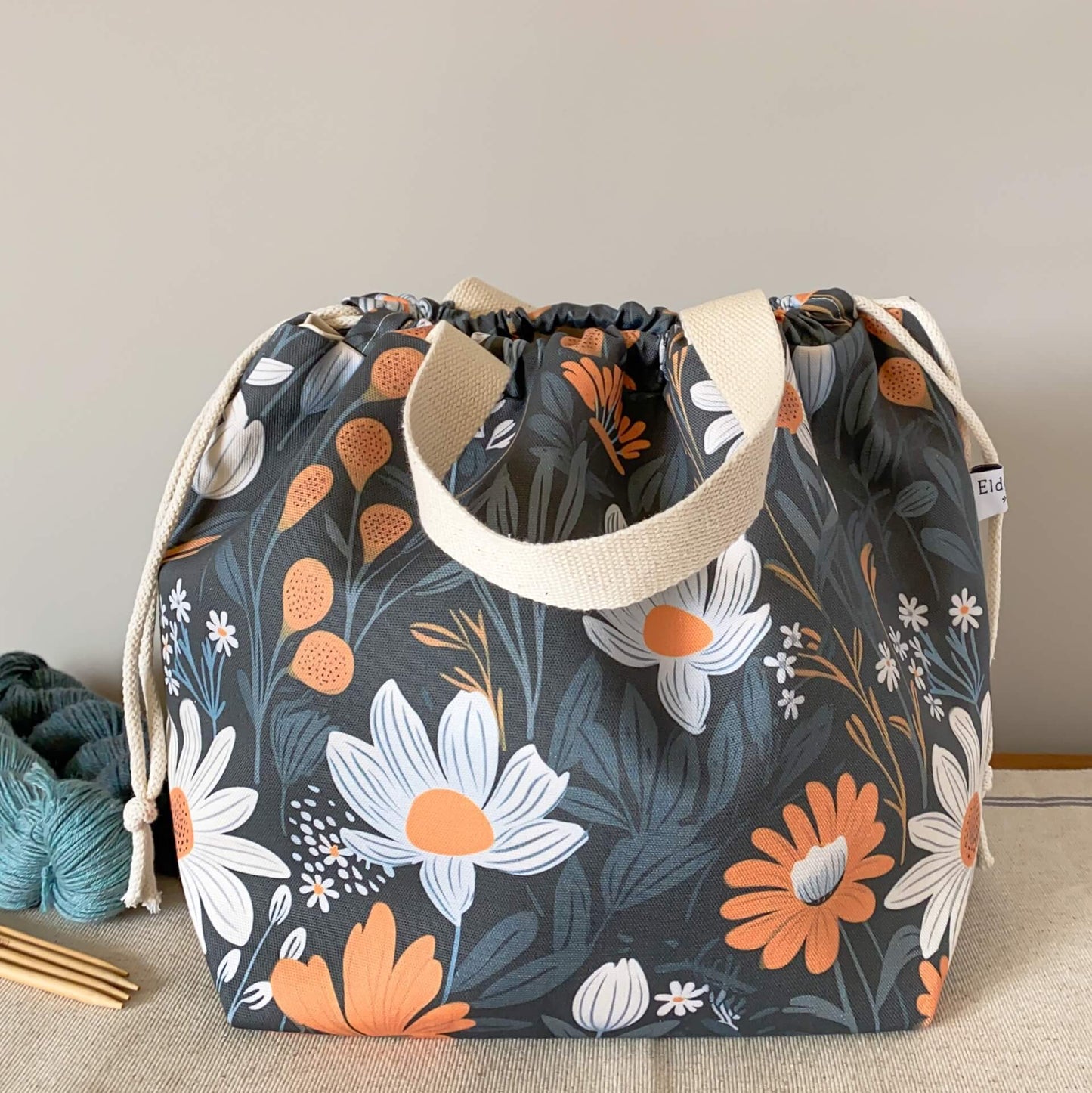 A knitting project bag featuring a bold floral printed fabric sits on a wooden table. It is pulled closed by drawstrings hiding the contents of the bag. 