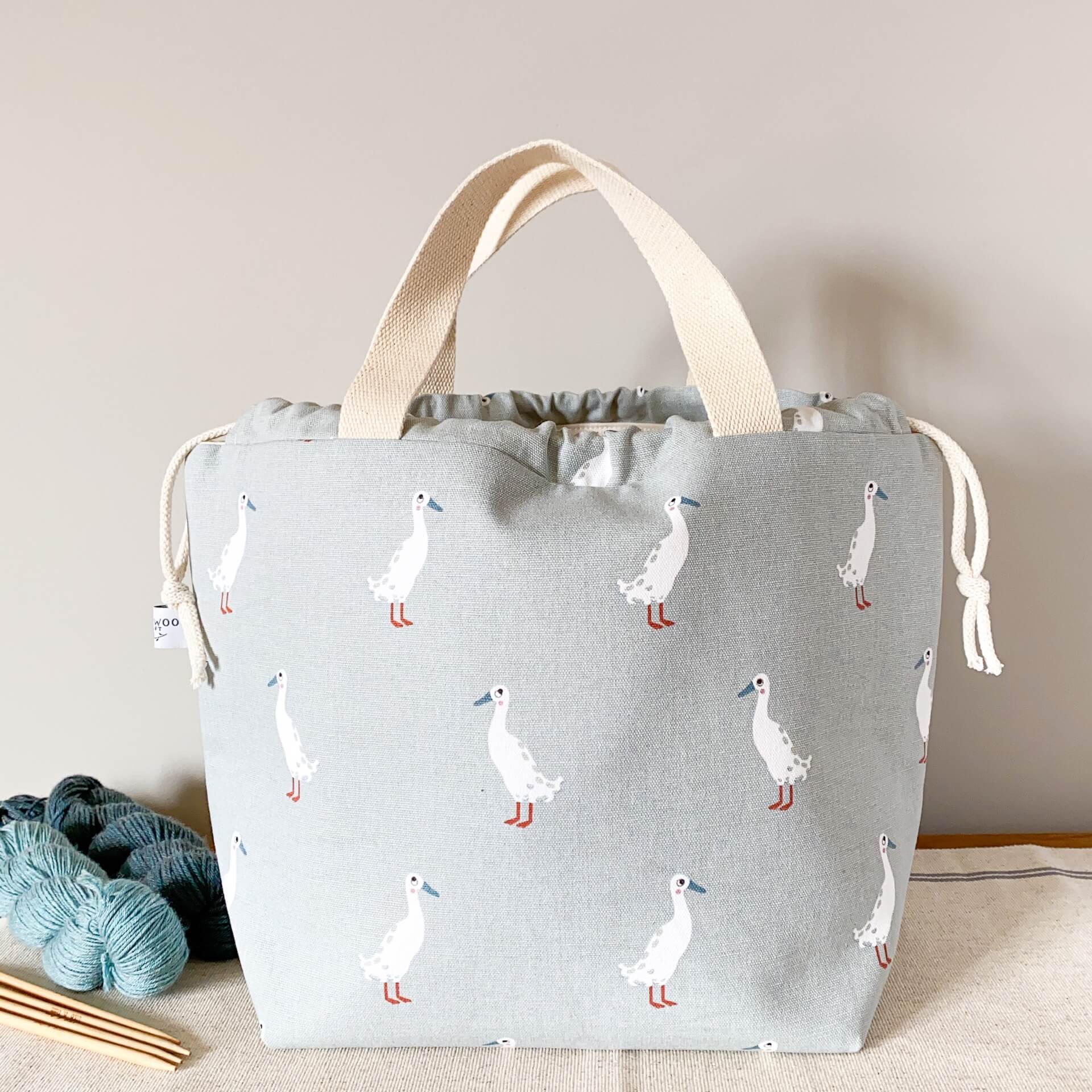 A large duck printed fabric bag sits on a table next to two skeins of yarn and knitting needles. The bag  has a grey background and the main feature of the fabric is some white runner ducks. 