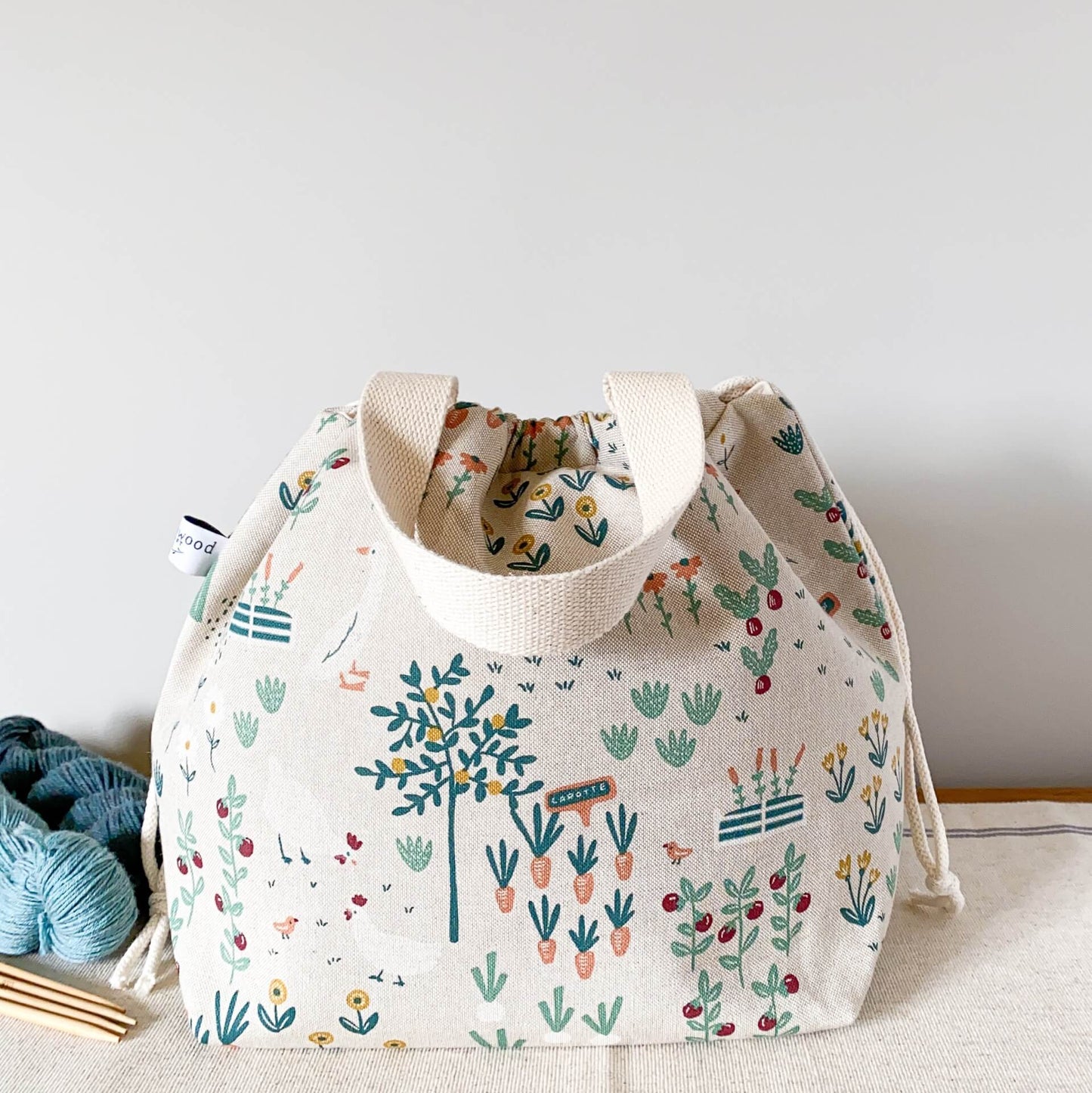 A cottage garden themed fabric sits on a table next to some yarn and knitting needles. The bag is pulled closed hiding its contents. 