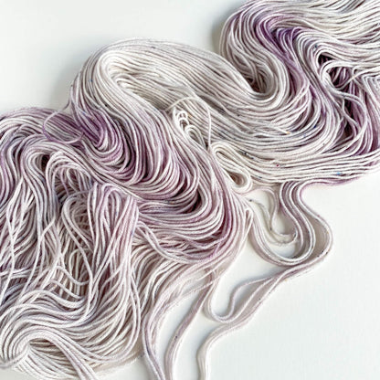 Hand dyed yarn lies scattered across a white table top. The yarn has been dyed with purple and grey dyes. 