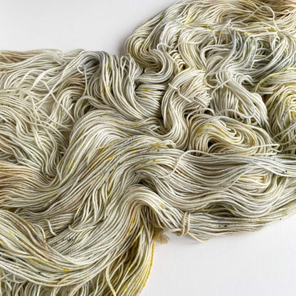 Hand dyed yarn unfurled from its skein has been tossed onto a white table top. The yarn shows colours of brown, grey and yellow. 